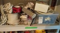 Photo of assorted electrical wiring spools, rolls of electrical tape, power strip, and plastic container with electrical components on a shelf.
