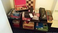 Overview image showing many boxed board and card games stacked on a small shelving unit, including a wooden chess set open with pieces inside, boxed dominoes, cards, and a large carved wooden box on top.