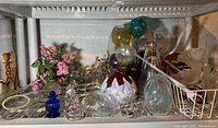 Photo of assorted glass decorative items including multiple plant waterers, blue glass bottle, and glass sculptures on a white metal shelf.