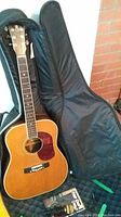 Full view of guitar leaning in open soft case, closed case behind, with microphone and songbook at base