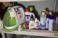 Bissell Littlegreen ProHeat Turbobrush spot cleaner with green hose and brush attachment, alongside multiple bottles of cleaning solutions on a shelf.