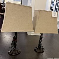 Two brown base table lamps with beige square tapered fabric shades, cords coiled at base, displayed on dark wooden floor.