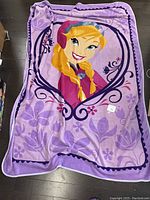 Photo showing full view of a purple blanket laid flat on floor featuring a blonde-haired character from Disney Frozen with floral designs and decorative border
