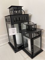 Three black metal lanterns with clear glass panels, one contains a white candle inside. Lanterns have vertical rectangular vents and top handles.