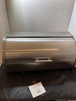 Metal bread box with roll-top lid and handle shown on black surface with white wall background.