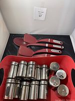Photo showing 10 stainless steel exterior glass spice jars arranged in two rows on a red tray, along with three red KitchenAid spatulas and one ladle with black and stainless steel accented handles.