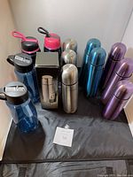 Lot of various thermoses and water bottles displayed against a white background on a dark mat, including stainless steel thermoses and plastic water bottles.