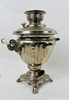 Front angled view of the silver-tone vintage samovar showing its ornate handles, lid with white knob, and base with feet.