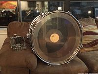 Bass drum with clear front drumhead and pedal to the left on a brown couch in front of window.