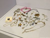 Lot of assorted jewelry chains and pendants laid out on a white surface, showing variety of materials, colors, and styles.