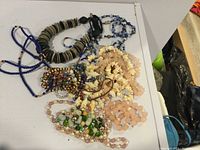 Full view of assorted beaded necklaces in various colors and materials including stone, glass, rose quartz, and shell beads placed on white surface.