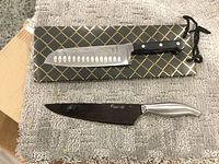 Two chef kitchen knives shown on carpet, one EarthChef BergHOFF with black handle, hollow edge blade; one Presidents Choice with silver handle and black blade.