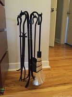 Wrought iron fireplace tool set with four tools and stand on a hardwood floor near white wall.