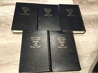 Five hardcover books with black covers and gold embossed Hebrew and English titles laid flat on wooden floor.