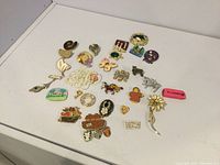 Photo showing 27 assorted pins and brooches laid out on a white surface, highlighting varied materials including enameled metal, wood, beads, painted glass, and rhinestones.