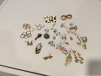 Full view of various rings and earrings arranged on white surface showing assortment and some needing repair.