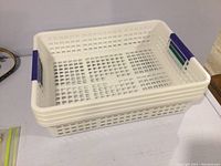 Four white plastic storage baskets stacked, showing purple handles and vented grid pattern on the sides and bottom.