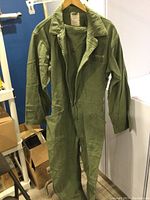 Full view of the vintage US Navy khaki green coveralls hanging on a wooden hanger showing front side and detail of pockets and collar.
