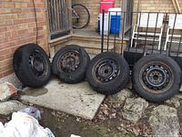 Four winter tires mounted on steel rims lined up outside against a brick wall. Tires appear to be in good condition while rims show rust and need cleaning.