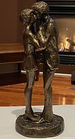 Front side of bronze loving couple sculpture showing intimate embrace and kiss with detailed clothing and textured hair, set against indoor background with fireplace lighting.
