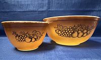 Two Pyrex mixing bowls showing the yellow exterior with black grape and apple fruit pattern detail in Mid-Century Modern style.