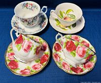 Four vintage teacup and saucer sets displayed on blue fabric, showing floral and fruit designs with gold trim.
