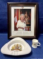Framed photo print of Queen Elizabeth II with milk glass ashtray and porcelain mug placed in front on blue fabric background.