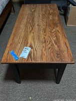 Top and side view of rectangular wooden coffee table showing brown wood grain top and black painted legs with two drawer fronts.