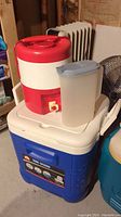 Blue and white Ice Cube cooler with red and white insulated drink dispenser and plastic water jug on top.