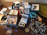 Photo showing a collection of various costume jewelry items including earrings, pins, necklaces, and bracelets spread on a table, some in boxes and original packaging.