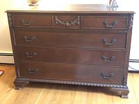 Full frontal view of the dresser showing the four drawers with detailed wood carving and metal handles, on wood flooring with heating baseboard behind.