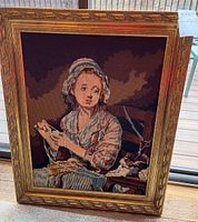 Full view of the framed needlepoint artwork showing the woman wrapping yarn and the kitten, with detailed ornate gold-painted wooden frame, standing on a wooden deck.