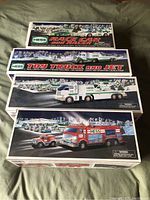 Four Hess toy trucks in original boxes displayed on a fabric surface. Boxes have winter scene backgrounds and images of trucks paired with smaller vehicles or jets.