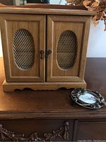 Solid wood musical jewelry box with two front doors, each having an oval metal mesh insert and antique-style metal handles. The box has scalloped base trim and an oak finish.