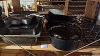 Photo of multiple kitchen items including cast iron Dutch oven with glass lid, wok, roasting racks, and roasting pans on wooden shelving