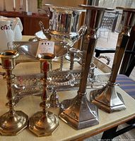Photo showing six candlesticks (four tall with square bases, two shorter with hexagonal bases) and an ice bucket, metal trays and stands on table.