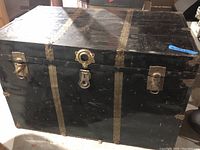 Front view of flat top metal travel steamer trunk with brass-tone vertical straps and lock center with two side latches. Displays visible wear and scratches on painted surface.