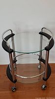 Full view of the round tempered glass bar cart showing two glass shelves with metal and black wood frame supports on casters.