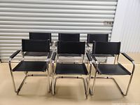 Front view of six black dining chairs with chrome metal tubular frames and black leather seats, backs, and armrests.