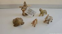 Photo shows five hand carved items arranged on white surface: turtle on left front, small dog figure and elephant on right front, bear behind elephant, and Inuksuk stone figure at back center with a descriptive card.