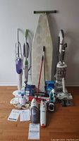 Multiple floor care items including Vileda sweeper, H2O steam mop, Bissell steam mop with pads, H2O hand steamer, Shark vacuum, B&D dustbusters, iron and ironing board