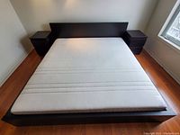 Wide shot of assembled dark brown IKEA king size bed frame with white mattress installed. Includes headboard with bedside drawer structures.