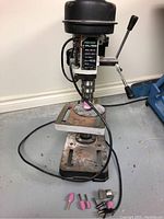 Rexon 8-inch drill press on floor with several grinding and wire brush bits nearby.