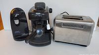 Three small kitchen appliances on a white surface: Black & Decker can opener, DeLonghi Caffe Parma espresso machine, and a Hamilton Beach toaster.