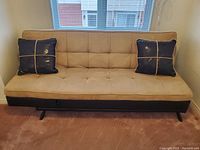 Front view of three-seat tan fabric futon sofa with two dark blue throw pillows, showing button tufting and black base.