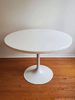 Full view of the white tulip table against a white wall on wooden floor, showing round top and pedestal base.