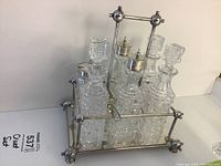 Front view of the six-piece glass cruet set arranged in the metal tray and holder with handle. Four bottles have glass stoppers; two have metal shaker tops.