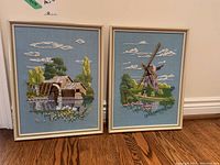 Two framed embroidery pieces side by side showing water mill and windmill scenes on blue fabric.