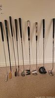 Full length view showing 9 golf club putters with different head and grip styles lined up on carpeted floor against white wall
