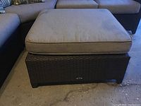 Front view of the Hampton Bay wicker ottoman with beige cushioned top on concrete floor.